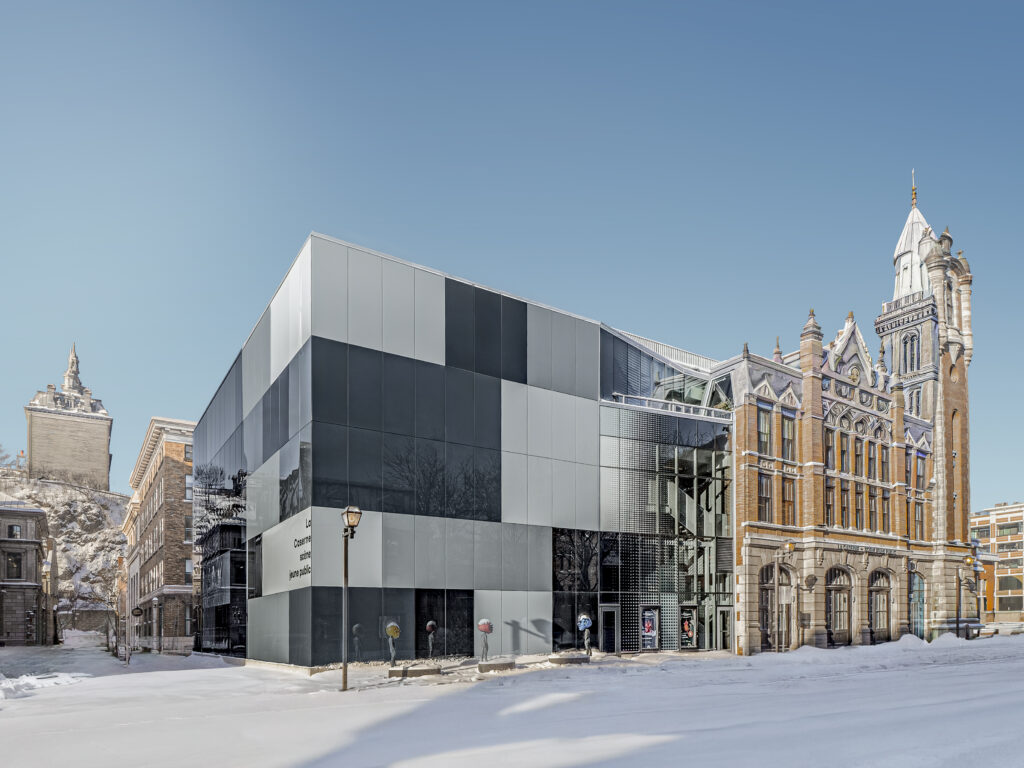 Façade vitrée réfléchissante du Théâtre des Gros Becs à Québec, avec panneaux en verre opaque et reflets des bâtiments voisins.